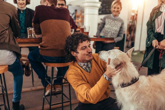 A man pets a dog while people sit at a bar behind him