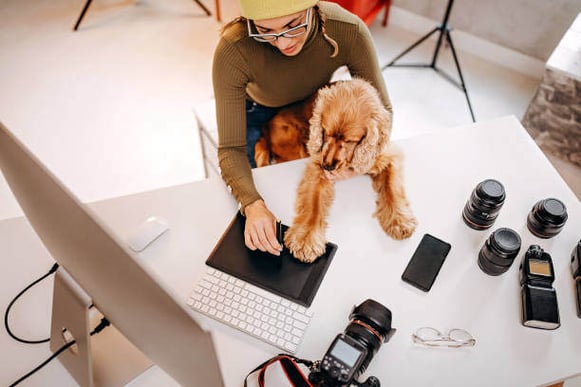 A woman uses a computer as a dog sits on her lap, with camera equipment laying on the desk in front of them