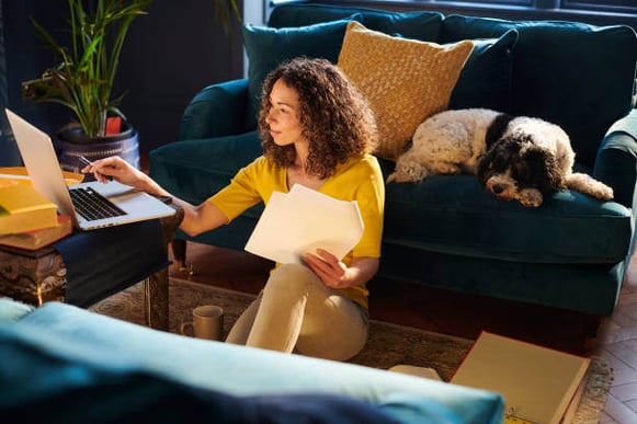 A woman sits on the floor using a laptop as her dog sits on the couch behind her