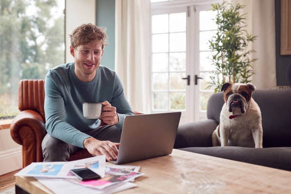 A man drinks coffee while using a laptop, as a dog sits in a chair next to him