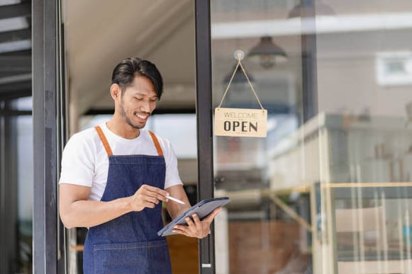 A man wearing an apron uses an ipad in front of a store with an open sign on the window