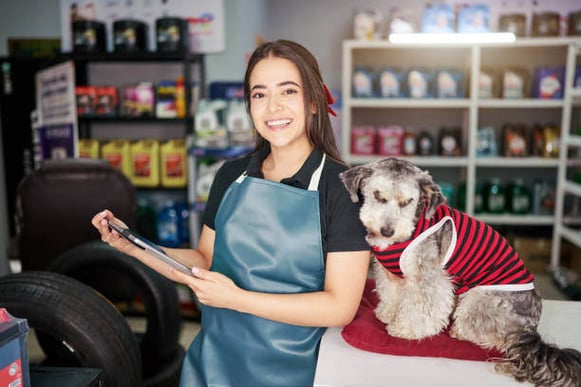 A dog groomer holds a clipboard standing next to a dog on a grooming table